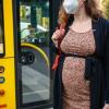 Eine schwangere Frau wartet mit Mund-Nasen-Schutz an einer Haltestelle auf dem Bus.