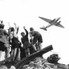 West-Berliner Jungen, die auf einem Trümmerberg stehen, begrüßen winkend ein US-amerikanisches Transportflugzeug, das Versorgungsgüter nach West-Berlin bringt. 