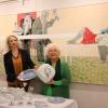 Die Künstlerin Anneliese Neumann und ihre Laudatorin Astrid Thum präsentieren vor einer dreidimensionalen Tafel von Hand bemaltes Geschirr. 