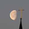 Der Mond steht im Morgenlicht hinter einem Kreuz auf einem Kirchturm.