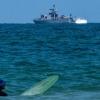 Ein Surfer wartet auf eine Welle, während ein israelisches Marineschiff im Mittelmeer vor der Küste von Hadera patrouilliert.