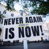 Ein Schild mit der Aufschrift „Never again is now!“ ist bei Teilnehmern einer Mahnwache gegen ein geplantes Pro-Palästina-Protestcamp vor der Ludwig-Maximilians-Universität (LMU) zu sehen.
