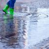 Mit Gummistiefeln geht eine Frau über eine leicht überflutete Straße.