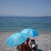 Eine Frau sitzt mit einem Handventilator unter einem Sonnenschirm am Strand in Griechenland. Urlauber auf Kreta sollten bei der Hitze vorsichtig sein.