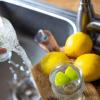 Tinkwasser läuft aus dem Wasserhahn in einer Küche in ein Glas mit Limettenstücken, während im Vordergrund Zitronen auf einem Küchenbrett liegen.