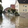 Um kurz nach 5 Uhr, am Tag der Deutschen Einheit, ist die Mindel in Jettingen (Landkreis Günzburg) übergelaufen und hat ein Wohngebiet überschwemmt. Offenbar war eine sogenannte Falle am Wasserkraftwerk defekt. Durch den starken Regen konnte nicht mehr genügend Wasser abfließen.

Wie Einsatzleiter und Kommandant der örtlichen Feuerwehr Markus Schmucker mitteilte, stand im "Korea", wie die Einheimischen das Wohngebiet am westlichen Ortsrand von Jettingen nennen, das Wasser auf den Straßen rund 20 Zentimeter hoch, als die Einsatzkräfte kamen. Und das Wasser stieg noch weiter.
Schleuse müssen geöffnet werdenAm ehemaligen Stauffenberg-Areal zwischen Mindellauf und Mindelkanal befindet sich ein Wasserkraftwerk mit zwei großen Fallen. Bei geringer ankommender Wassermenge sind beide Fallen geschlossen, um das Wasser komplett durch die Turbine zur Stromerzeugung zu leiten. Kommt aber eine so große Wassermenge von flussaufwärts an, dass das Wasser durch den alleinigen Durchfluss durch die Turbine nicht mehr abfließen kann, müssen die Fallen geöffnet werden, um ein Überlaufen der Mindel zu verhindern.Defekt an einer der SchleusenDie zweite Falle wird geöffnet, wenn notwendig. Wie der 2. Bürgermeister von Jettingen-Scheppach Hans Reichhardt berichtet, funktioniert diese zweite Falle seit geraumer Zeit allerdings nicht. Das Gestänge ist ausgehängt. Die Regenmengen am Montag waren so stark, dass das vom Oberlauf der Mindel ankommende Wasser nicht mehr genügend abgeleitet werden konnte. Das führte zum Ansteigen und schließlich zum Überlaufen der Mindel.Mindel läuft über!Das Wasser floss über mehrere Felder und überflutete schließlich das Wohngebiet bis zu einer Höhe von 1,20 Metern. Wie Reichhardt, der selbst ein Wasserkraftwerk betreibt, weiter ausführt, hatten Anwohner und die Gemeinde die Kraftwerksbetreiberin, die nicht im Landkreis wohnt, bereits dazu aufgefordert, die defekte Falle instand zu setzen. Dies sei leider bisher nicht geschehen, mit den nun entsprechenden Konsequenzen. Letztlich wurde ein von der Gemeinde betriebener Kanal geöffnet, über den eine große Wassermenge um das Wohngebiet herum abgeleitet werden kann.Große Schäden in PrivathäusernDie Familie Wolfmiller wohnt im Hammerschmiedweg und wurde vom Nachbarn gegen 6 Uhr angerufen und auf das Wasser aufmerksam gemacht. Wie der Vater sichtlich mitgenommen erklärt, hatte er dann mit einem Wassersauger im Keller das bereits dort angesammelte Wasser abgesaugt, als er plötzlich in einem Nebenraum ein Rauschen wahrnahm. Beim Nachschauen konnte er feststellen, dass dort die Bodenplatte des betonierten Kellers aufriss und das Wasser den Keller flutete. Neben Waschmaschine, Trockner und zwei Gefriertruhen ist auch der Speicher der Solaranlage im Keller. Alle Gerätschaften werden wohl komplett beschädigt worden sein.80 Kräfte im EinsatzZwischenzeitlich hatte der Einsatzleiter Schmucker u.a. noch die Feuerwehren Scheppach und Burgau nachalarmieren lassen. Insgesamt waren rund 80 Feuerwehrkräfte im Einsatz. Sie halfen mit rund 20 Tauch- und Tragkraftpumpen den Anwohnern beim vorsichtigen Auspumpen der Räumlichkeiten. Hier mussten die Einsatzkräfte aufpassen: Wenn der Wasserspiegel außen noch zu hoch ist, der Keller aber komplett leer gepumpt wird, wirkt ein großer Druck auf die Außenwände und die Bodenplatte eines Gebäudes. Das kann zu großen Schäden führen.Noch nie in diesem Ausmaß
Monika Wiesbauer, in deren Keller ebenfalls Wasser stand, zeigte ihren Garten. Dort hatte sie  noch am Sonntag Holz aufgestapelt und Blumen gerichtet. Nun steht dort das Wasser mehrere Zentimeter hoch. Die 68-Jährige wohnt dort seit 1958 und hat schon einige Male miterlebt, wie das Wasser aus dem Boden in den Keller gedrückt wurde. Dass es aber von oben in den Keller läuft und noch dazu in diesem Ausmaß, das gab es noch nie. Sie wurde gegen 6.30 Uhr von ihrem Nachbarn geweckt mit den Worten "Monika mach auf, da kommt 's Wasser." Doch viel dagegen tun konnte sie nicht. Hochwassergebiet ausgewiesenAuf die betroffenen Anwohner kommt eine schwere Zeit zu. Das Gebiet ist als Hochwassergebiet ausgewiesen. Daher ist es nicht möglich, ihre Häuser gegen solche Schäden zu versichern. Zudem müssen sie nun versuchen, die wassergetränkten Wände wieder trocken zu bekommen, was bei den aktuellen und weiter steigenden Energiekosten nochmals eine große Herausforderung darstellt. Ganz abgesehen von den beschädigten Gerätschaften und Heizungsanlagen.