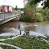 Um kurz nach 5 Uhr, am Tag der Deutschen Einheit, ist die Mindel in Jettingen (Landkreis Günzburg) übergelaufen und hat ein Wohngebiet überschwemmt. Offenbar war eine sogenannte Falle am Wasserkraftwerk defekt. Durch den starken Regen konnte nicht mehr genügend Wasser abfließen.

Wie Einsatzleiter und Kommandant der örtlichen Feuerwehr Markus Schmucker mitteilte, stand im "Korea", wie die Einheimischen das Wohngebiet am westlichen Ortsrand von Jettingen nennen, das Wasser auf den Straßen rund 20 Zentimeter hoch, als die Einsatzkräfte kamen. Und das Wasser stieg noch weiter.
Schleuse müssen geöffnet werdenAm ehemaligen Stauffenberg-Areal zwischen Mindellauf und Mindelkanal befindet sich ein Wasserkraftwerk mit zwei großen Fallen. Bei geringer ankommender Wassermenge sind beide Fallen geschlossen, um das Wasser komplett durch die Turbine zur Stromerzeugung zu leiten. Kommt aber eine so große Wassermenge von flussaufwärts an, dass das Wasser durch den alleinigen Durchfluss durch die Turbine nicht mehr abfließen kann, müssen die Fallen geöffnet werden, um ein Überlaufen der Mindel zu verhindern.Defekt an einer der SchleusenDie zweite Falle wird geöffnet, wenn notwendig. Wie der 2. Bürgermeister von Jettingen-Scheppach Hans Reichhardt berichtet, funktioniert diese zweite Falle seit geraumer Zeit allerdings nicht. Das Gestänge ist ausgehängt. Die Regenmengen am Montag waren so stark, dass das vom Oberlauf der Mindel ankommende Wasser nicht mehr genügend abgeleitet werden konnte. Das führte zum Ansteigen und schließlich zum Überlaufen der Mindel.Mindel läuft über!Das Wasser floss über mehrere Felder und überflutete schließlich das Wohngebiet bis zu einer Höhe von 1,20 Metern. Wie Reichhardt, der selbst ein Wasserkraftwerk betreibt, weiter ausführt, hatten Anwohner und die Gemeinde die Kraftwerksbetreiberin, die nicht im Landkreis wohnt, bereits dazu aufgefordert, die defekte Falle instand zu setzen. Dies sei leider bisher nicht geschehen, mit den nun entsprechenden Konsequenzen. Letztlich wurde ein von der Gemeinde betriebener Kanal geöffnet, über den eine große Wassermenge um das Wohngebiet herum abgeleitet werden kann.Große Schäden in PrivathäusernDie Familie Wolfmiller wohnt im Hammerschmiedweg und wurde vom Nachbarn gegen 6 Uhr angerufen und auf das Wasser aufmerksam gemacht. Wie der Vater sichtlich mitgenommen erklärt, hatte er dann mit einem Wassersauger im Keller das bereits dort angesammelte Wasser abgesaugt, als er plötzlich in einem Nebenraum ein Rauschen wahrnahm. Beim Nachschauen konnte er feststellen, dass dort die Bodenplatte des betonierten Kellers aufriss und das Wasser den Keller flutete. Neben Waschmaschine, Trockner und zwei Gefriertruhen ist auch der Speicher der Solaranlage im Keller. Alle Gerätschaften werden wohl komplett beschädigt worden sein.80 Kräfte im EinsatzZwischenzeitlich hatte der Einsatzleiter Schmucker u.a. noch die Feuerwehren Scheppach und Burgau nachalarmieren lassen. Insgesamt waren rund 80 Feuerwehrkräfte im Einsatz. Sie halfen mit rund 20 Tauch- und Tragkraftpumpen den Anwohnern beim vorsichtigen Auspumpen der Räumlichkeiten. Hier mussten die Einsatzkräfte aufpassen: Wenn der Wasserspiegel außen noch zu hoch ist, der Keller aber komplett leer gepumpt wird, wirkt ein großer Druck auf die Außenwände und die Bodenplatte eines Gebäudes. Das kann zu großen Schäden führen.Noch nie in diesem Ausmaß
Monika Wiesbauer, in deren Keller ebenfalls Wasser stand, zeigte ihren Garten. Dort hatte sie  noch am Sonntag Holz aufgestapelt und Blumen gerichtet. Nun steht dort das Wasser mehrere Zentimeter hoch. Die 68-Jährige wohnt dort seit 1958 und hat schon einige Male miterlebt, wie das Wasser aus dem Boden in den Keller gedrückt wurde. Dass es aber von oben in den Keller läuft und noch dazu in diesem Ausmaß, das gab es noch nie. Sie wurde gegen 6.30 Uhr von ihrem Nachbarn geweckt mit den Worten "Monika mach auf, da kommt 's Wasser." Doch viel dagegen tun konnte sie nicht. Hochwassergebiet ausgewiesenAuf die betroffenen Anwohner kommt eine schwere Zeit zu. Das Gebiet ist als Hochwassergebiet ausgewiesen. Daher ist es nicht möglich, ihre Häuser gegen solche Schäden zu versichern. Zudem müssen sie nun versuchen, die wassergetränkten Wände wieder trocken zu bekommen, was bei den aktuellen und weiter steigenden Energiekosten nochmals eine große Herausforderung darstellt. Ganz abgesehen von den beschädigten Gerätschaften und Heizungsanlagen.