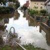 Um kurz nach 5 Uhr, am Tag der Deutschen Einheit, ist die Mindel in Jettingen (Landkreis Günzburg) übergelaufen und hat ein Wohngebiet überschwemmt. Offenbar war eine sogenannte Falle am Wasserkraftwerk defekt. Durch den starken Regen konnte nicht mehr genügend Wasser abfließen.

Wie Einsatzleiter und Kommandant der örtlichen Feuerwehr Markus Schmucker mitteilte, stand im "Korea", wie die Einheimischen das Wohngebiet am westlichen Ortsrand von Jettingen nennen, das Wasser auf den Straßen rund 20 Zentimeter hoch, als die Einsatzkräfte kamen. Und das Wasser stieg noch weiter.
Schleuse müssen geöffnet werdenAm ehemaligen Stauffenberg-Areal zwischen Mindellauf und Mindelkanal befindet sich ein Wasserkraftwerk mit zwei großen Fallen. Bei geringer ankommender Wassermenge sind beide Fallen geschlossen, um das Wasser komplett durch die Turbine zur Stromerzeugung zu leiten. Kommt aber eine so große Wassermenge von flussaufwärts an, dass das Wasser durch den alleinigen Durchfluss durch die Turbine nicht mehr abfließen kann, müssen die Fallen geöffnet werden, um ein Überlaufen der Mindel zu verhindern.Defekt an einer der SchleusenDie zweite Falle wird geöffnet, wenn notwendig. Wie der 2. Bürgermeister von Jettingen-Scheppach Hans Reichhardt berichtet, funktioniert diese zweite Falle seit geraumer Zeit allerdings nicht. Das Gestänge ist ausgehängt. Die Regenmengen am Montag waren so stark, dass das vom Oberlauf der Mindel ankommende Wasser nicht mehr genügend abgeleitet werden konnte. Das führte zum Ansteigen und schließlich zum Überlaufen der Mindel.Mindel läuft über!Das Wasser floss über mehrere Felder und überflutete schließlich das Wohngebiet bis zu einer Höhe von 1,20 Metern. Wie Reichhardt, der selbst ein Wasserkraftwerk betreibt, weiter ausführt, hatten Anwohner und die Gemeinde die Kraftwerksbetreiberin, die nicht im Landkreis wohnt, bereits dazu aufgefordert, die defekte Falle instand zu setzen. Dies sei leider bisher nicht geschehen, mit den nun entsprechenden Konsequenzen. Letztlich wurde ein von der Gemeinde betriebener Kanal geöffnet, über den eine große Wassermenge um das Wohngebiet herum abgeleitet werden kann.Große Schäden in PrivathäusernDie Familie Wolfmiller wohnt im Hammerschmiedweg und wurde vom Nachbarn gegen 6 Uhr angerufen und auf das Wasser aufmerksam gemacht. Wie der Vater sichtlich mitgenommen erklärt, hatte er dann mit einem Wassersauger im Keller das bereits dort angesammelte Wasser abgesaugt, als er plötzlich in einem Nebenraum ein Rauschen wahrnahm. Beim Nachschauen konnte er feststellen, dass dort die Bodenplatte des betonierten Kellers aufriss und das Wasser den Keller flutete. Neben Waschmaschine, Trockner und zwei Gefriertruhen ist auch der Speicher der Solaranlage im Keller. Alle Gerätschaften werden wohl komplett beschädigt worden sein.80 Kräfte im EinsatzZwischenzeitlich hatte der Einsatzleiter Schmucker u.a. noch die Feuerwehren Scheppach und Burgau nachalarmieren lassen. Insgesamt waren rund 80 Feuerwehrkräfte im Einsatz. Sie halfen mit rund 20 Tauch- und Tragkraftpumpen den Anwohnern beim vorsichtigen Auspumpen der Räumlichkeiten. Hier mussten die Einsatzkräfte aufpassen: Wenn der Wasserspiegel außen noch zu hoch ist, der Keller aber komplett leer gepumpt wird, wirkt ein großer Druck auf die Außenwände und die Bodenplatte eines Gebäudes. Das kann zu großen Schäden führen.Noch nie in diesem Ausmaß
Monika Wiesbauer, in deren Keller ebenfalls Wasser stand, zeigte ihren Garten. Dort hatte sie  noch am Sonntag Holz aufgestapelt und Blumen gerichtet. Nun steht dort das Wasser mehrere Zentimeter hoch. Die 68-Jährige wohnt dort seit 1958 und hat schon einige Male miterlebt, wie das Wasser aus dem Boden in den Keller gedrückt wurde. Dass es aber von oben in den Keller läuft und noch dazu in diesem Ausmaß, das gab es noch nie. Sie wurde gegen 6.30 Uhr von ihrem Nachbarn geweckt mit den Worten "Monika mach auf, da kommt 's Wasser." Doch viel dagegen tun konnte sie nicht. Hochwassergebiet ausgewiesenAuf die betroffenen Anwohner kommt eine schwere Zeit zu. Das Gebiet ist als Hochwassergebiet ausgewiesen. Daher ist es nicht möglich, ihre Häuser gegen solche Schäden zu versichern. Zudem müssen sie nun versuchen, die wassergetränkten Wände wieder trocken zu bekommen, was bei den aktuellen und weiter steigenden Energiekosten nochmals eine große Herausforderung darstellt. Ganz abgesehen von den beschädigten Gerätschaften und Heizungsanlagen.