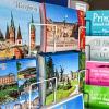 Wer kennt es nicht - das freudige Gefühl, wenn man den Briefkasten öffnet und von einem bunten Motiv aus einem fernen Land begrüßt wird? Postkarten mit einer persönlichen Nachricht aus dem Urlaub erfreuen auch heute noch die Familie und Freunde des Reisenden.   Aber werden heutzutage in den Zeiten von Handy und digitalen Netzwerken tatsächlich noch genau so viele Karten versendet wie noch vor dem  Beginn des digitalen Zeitalters? Spaziert man durch die Innenstadt Würzburgs, entdeckt man an auffallend vielen Ecken Verkaufsständer mit Postkarten, die die verschiedensten Sehenswürdigkeiten der Domstadt präsentieren. Als Einheimischer stellt man sich nun bisweilen die Frage: „Wie viele werden davon überhaupt noch verkauft?“   Werden Postkarten in Zeiten, in denen ein Selfie von der Alten Mainbrücke innerhalb von Sekunden bei den Lieben zu Hause ankommt, von Touristen eigentlich noch wahrgenommen? Eine Umfrage bei einigen Würzburger Läden gab Aufschluss.  Erste Anlaufstelle für Urlauber ist in den meisten Fällen der Dom. Der danebenliegende Shop „Volk - Kunst am Dom“ kann aufgrund dessen eine große Menge an verkauften Postkarten verbuchen. „Momentaner Trend sind Spruchkarten“, erzählt Ute Hut. Diese werden gleich samt Briefmarke gekauft und anschließend versendet. Antonia Pongratz von der Buchhandlung „Hugendubel“ bestätigt dies und kann von einer besonderen Vorliebe ihrer Käufer berichten: „Vor allem lustige Frankensprüche werden viel gekauft.“ Eine Ecke weiter, an der Marienkapelle, sieht Rita Herbert, die in der kleinen Filiale der „Max Jakob - Kerzenmanufaktur“ arbeitet, keine besondere Tendenz zu bestimmten Motiven: „Es wird kunterbunt gekauft, quer durchs Sortiment.“ Auf der anderen Seite der Kapelle, im Souvenir- und Geschenkshop „blumen piazza“, machen vor allem speziell für den Laden angefertigte Karten das Rennen.   „Die mit der Maria Empfängnis darauf wird am häufigsten gekauft“, berichtet der Inhaber. In Würzburg bekannt als „Die Jesusrutsche“, scheint das lokale Motiv durch seine Originalität zu überzeugen. Eine weitere Auffälligkeit wurde in allen vier Läden bemerkt: Der Großteil der Käufer sind junge Menschen. Allem Anschein nach lässt die Beliebtheit der Postkarte also trotz SMS und Internet selbst beim jungen Volk nicht nach. Doch wie sieht die Realität aus? Schreiben Würzburgs Touristen wirklich so viele Postkarten? Katharina Kaul ist für einen Tagestrip mit ihrer Familie in Würzburg. Auf den Stufen des Doms berichtet die junge Coburgerin: „Wenn wir länger in den Urlaub fahren, dann schreiben wir mindestens zehn bis fünfzehn Postkarten an Freunde und Familie. Am meisten freut sich unsere Nachbarin, eine ältere Dame, über unsere Post.“  Ebenfalls auf den Stufen des Würzburger Wahrzeichens sitzt eine französische Touristin. Brigitte Cassard zeigt stolz die neun Postkarten, die sie gerade erstanden hat. Auf der Weiterreise wird sie im Zug an ihre Familie und Freunde schreiben. Besonders beeindruckt ist sie von „so viel Auswahl hier in der Stadt“.  Eine Pressemitteilung der Deutschen Post untermauert die Kauffreudigkeit der Touristen: Jedes Jahr versenden sie alleine zwischen Juni und August mit der Deutschen Post circa 60 Millionen Ansichtskarten. Eine Umfrage, die kürzlich vom Tourismusunternehmen L'TUR durchgeführt wurde, hat gezeigt, dass 78 Prozent der Befragten noch immer eher dazu tendieren eine Postkarte zu verschicken anstatt zum Handy zu greifen.   Während 71 Prozent der Männer weiterhin den schriftlichen Urlaubsgruß versenden, überwiegt der weibliche Anteil mit 84 Prozent.
