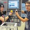 Einmal aus dem 3D-Drucker, einmal aus der CNC-Drehmaschine:  Jennifer Büttner hält den „gedruckten“ Becher aus Plastik, Yassin Bel Mahdi den „Zwillingsbruder“ aus Metall, der in der CNC-Drehmaschine (im Hintergrund) hergestellt wurde.