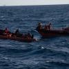 Migranten sitzen in einem Boot im Mittelmeer; Rettungskräfte versuchen, ihnen zu helfen.